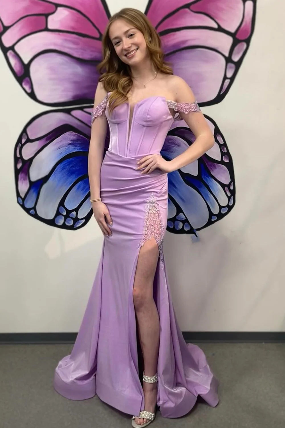 Model wearing a purple mermaid off-shoulder prom dress with applique and corset details, showcasing the thigh-high slit.