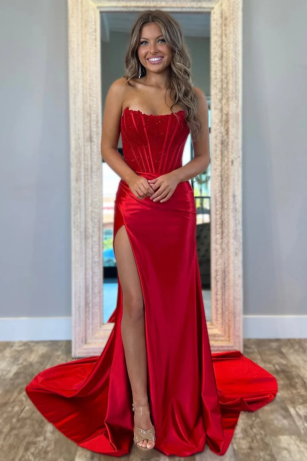 Woman wearing a long red strapless prom dress with a glitter top and a high leg slit.